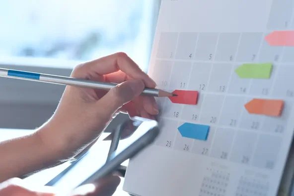 Hand markiert ein Datum auf einem Kalender mit bunten Markierungen – Symbol für den Wahlkalender der Betriebsratswahl.