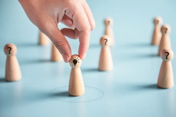 Hand wählt eine Spielfigur mit einer Glühbirne aus einer Gruppe mit Fragezeichen – Symbol für das Gewinnen von Kandidaten bei der Betriebsratswahl.